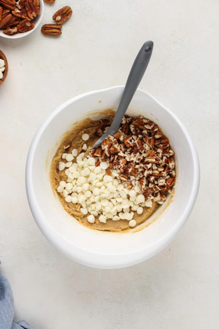 White chocolate chips and chopped pecans being added to blondie batter in a white bowl.