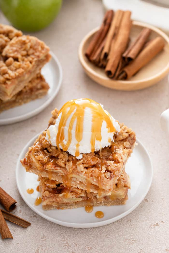 Stack of apple crisp bars topped with ice cream and caramel sauce on a white plate.