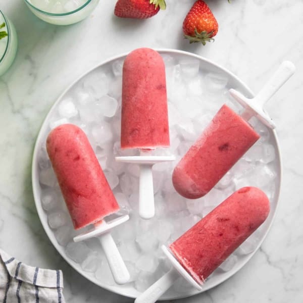 Four strawberry popsicles arranged on a bowl of ice on a marble countertop.