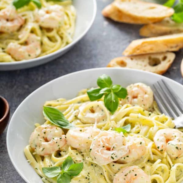 Plated bowl of creamy pesto pasta with shrimp with another bowl and slices of baguette in the background