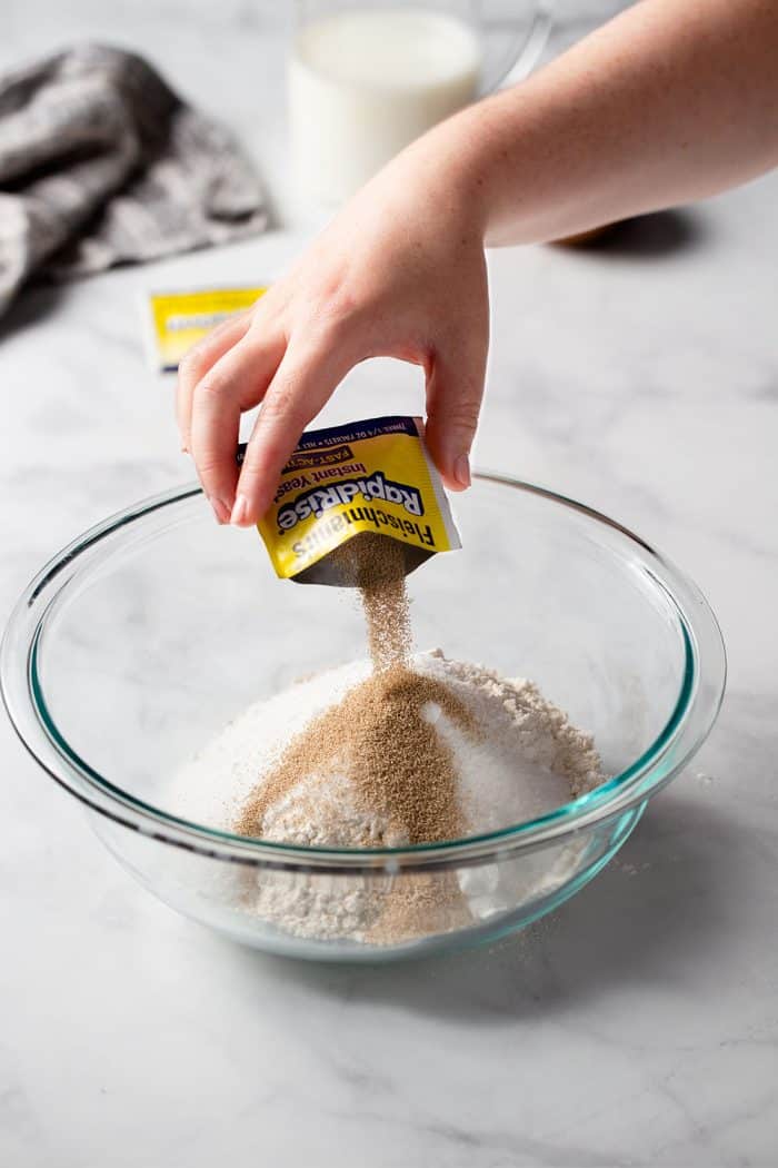 Hand pouring a packet of yeast into a glass mixing bowl with flour for parker house rolls