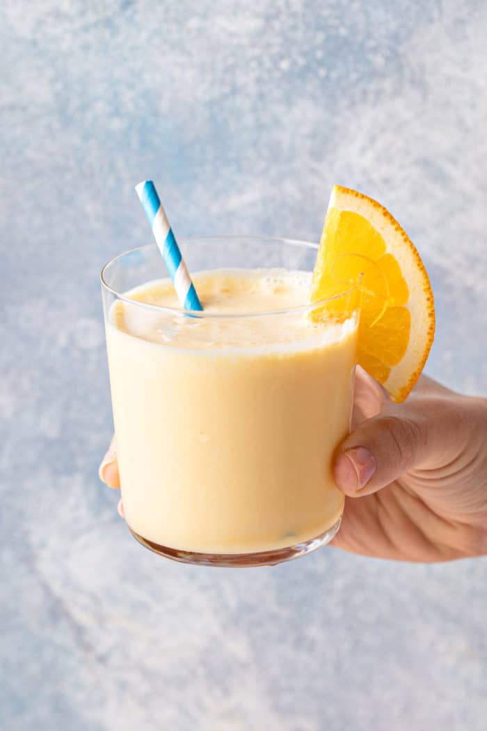 Hand holding up a glass of orange julius, garnished with a blue and white striped straw and an orange slice.