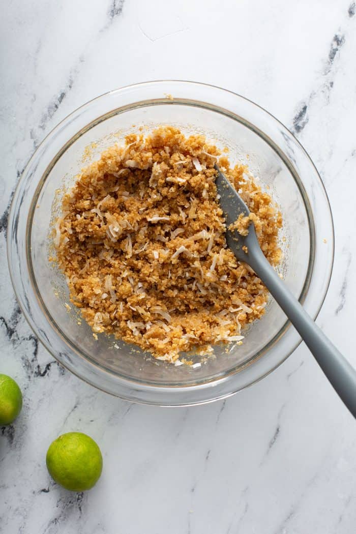 coconut graham cracker crust being mixed together with a spatula in a glass mixing bowl on a marble surface