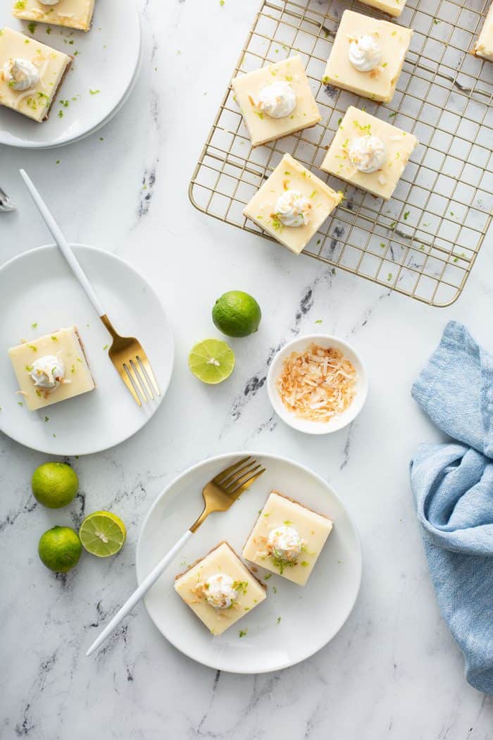 Overhead view of three white plates holding key lime pie bars on a marble counter next to a wire cooling rack holding more key lime pie bars