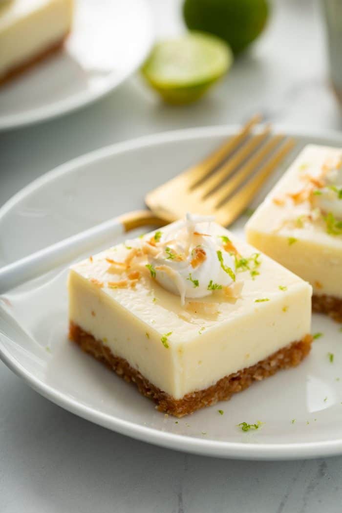 Close up of two key lime pie bars topped with whipped cream and toasted coconut on a white plate, next to a gold fork with a white handle