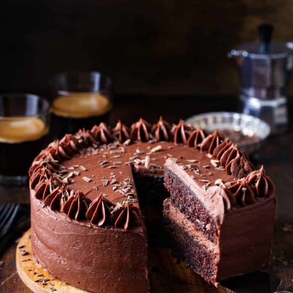 Slice being removed from a double layer chocolate cake with chocolate frosting, with coffee in the background