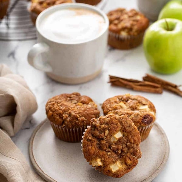 Three apple cinnamon muffins on a cream plate with lattes and more muffins on a cooling rack in the background