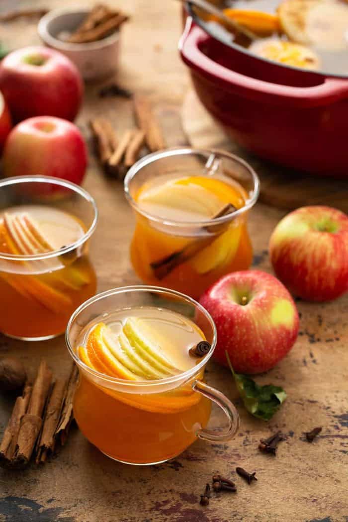 Three mugs of hot wassail garnished with sliced apples and cinnamon sticks with a dutch oven full of wassail in the background