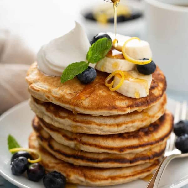 Stack of banana pancakes on a white plate, topped with sliced bananas, blueberries and whipped cream, with syrup being drizzled on top