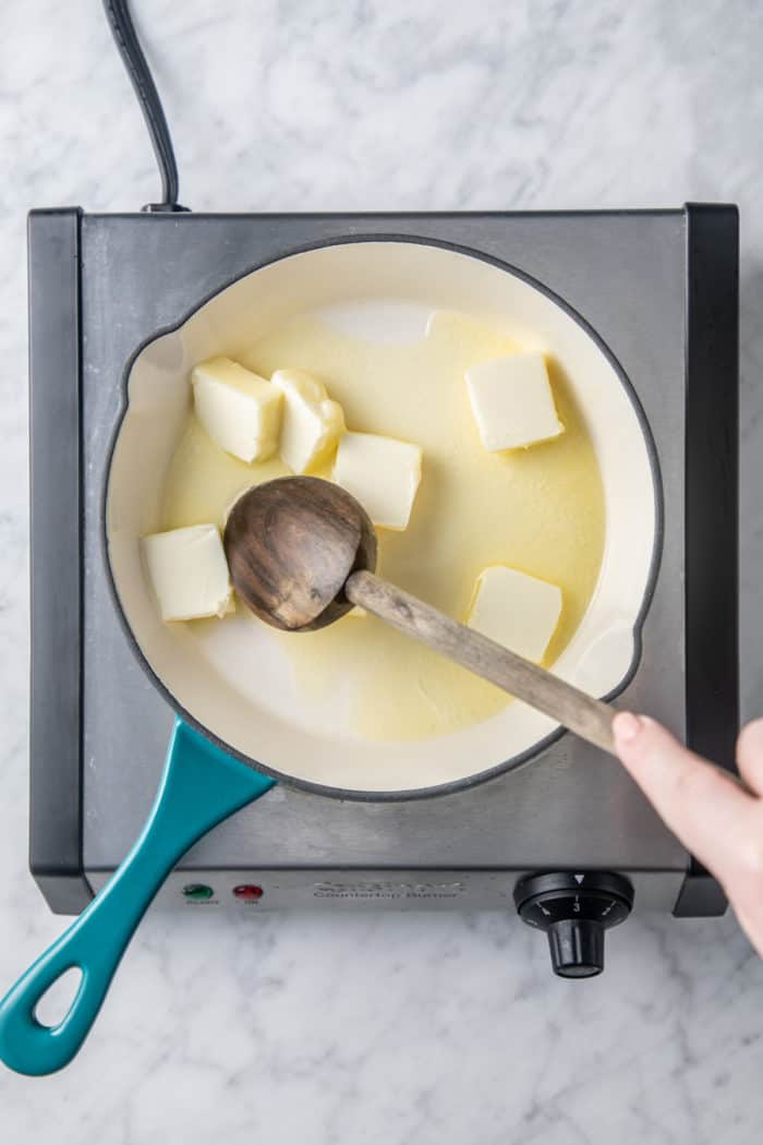 Pats of butter being melted in a light-colored skillet on a burner.