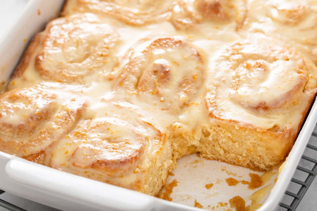 White baking dish of orange rolls with a roll removed from the corner of the pan.