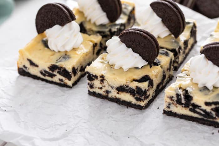 Sliced oreo cheesecake bars topped with whipped cream and more oreo cookies.