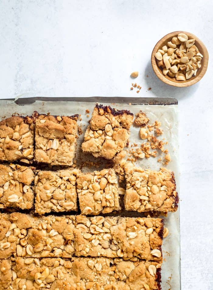 Sliced peanut butter and jelly bars on a parchment-lined tray next to a small bowl of roasted peanuts