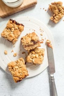 Four peanut butter and jelly bars scattered on a white plate next to a knife with peanut butter on the tip