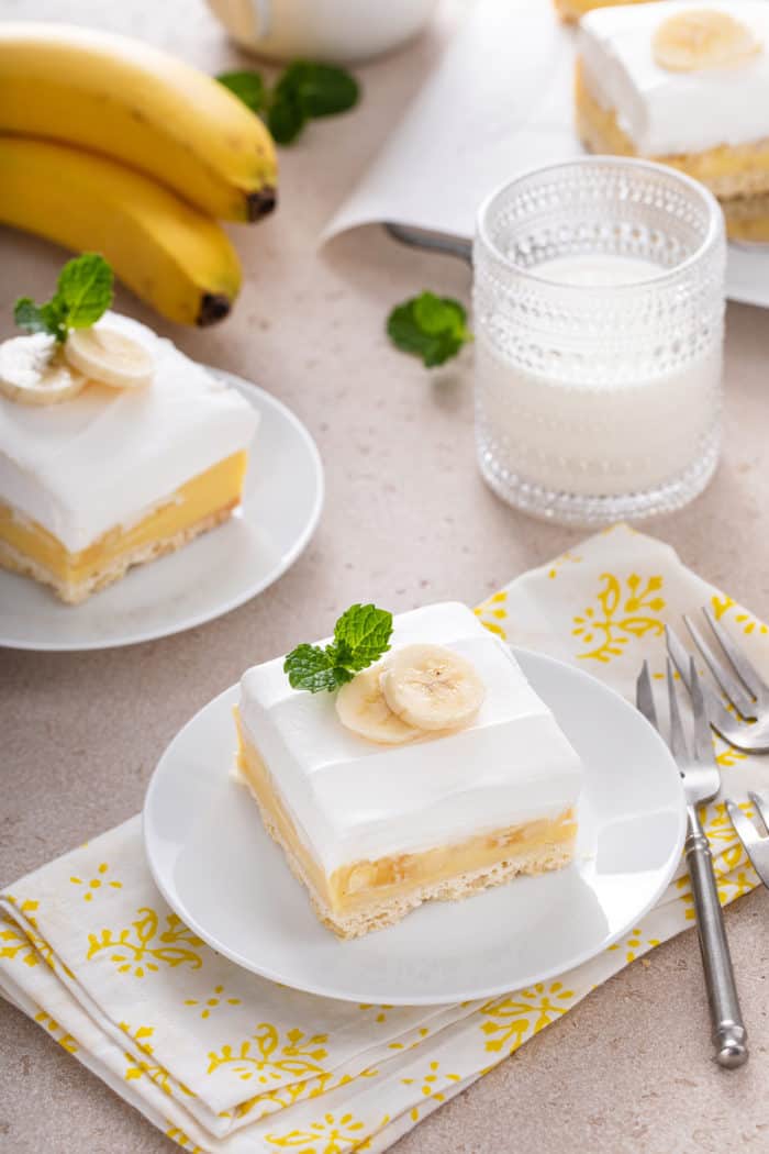 Multiple white plates, each with a banana cream bar on them. A glass of milk and a bunch of bananas are also visible in the shot.