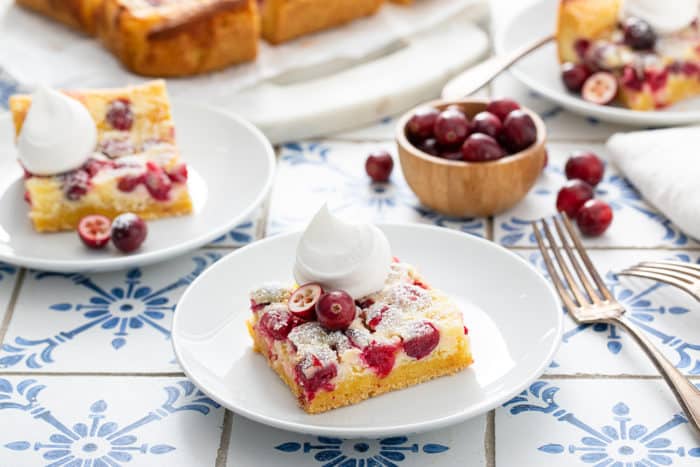 Two white plates with slices of gooey butter bars, each topped with whipped cream, set on a blue countertop