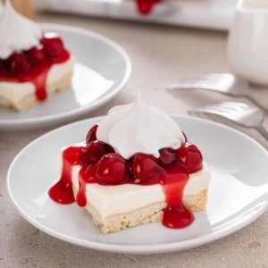 Slice of nana's easy cheesecake, topped with cherry pie filling and whipped cream, on a white plate.