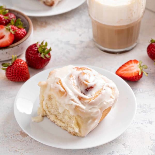 Strawberry roll with cream cheese frosting set on a white plate. Coffee and fresh strawberries are in the background.