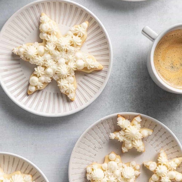 Assorted snowflake sugar cookies on white plates next to cups of coffee