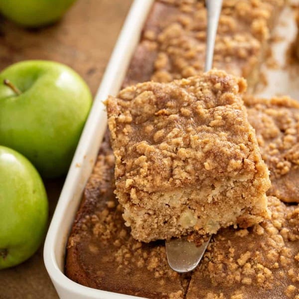 Cake server holding a slice of apple coffee cake over a pan of coffee cake