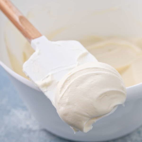 Spatula with cream cheese frosting on it set on the side of a white mixing bowl