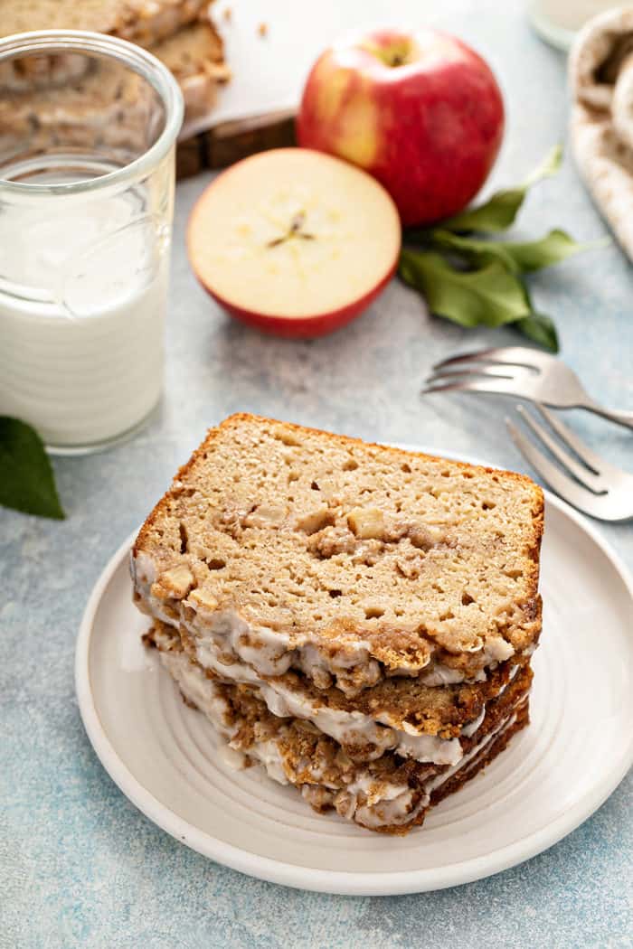 Three slices of apple fritter bread stacked on a white plate. A glass of milk and apples are in the background