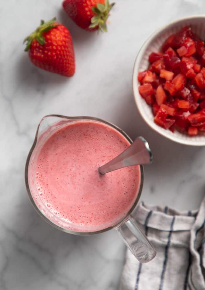 Strawberry popsicle base in a large glass measuring cup, next to a bowl of chopped strawberries.