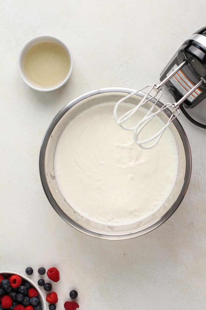 Hand mixer beating heavy cream in a metal bowl set next to a small bowl of melted unflavored gelatin.